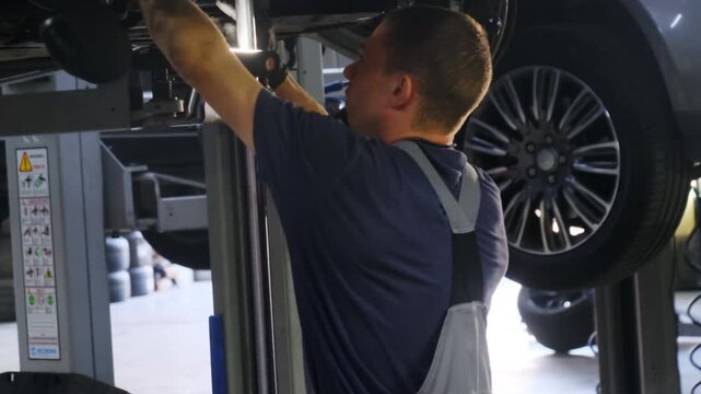 Technician uses flashlight to illuminate underside of vehicle. Man in overalls inspects parts on surface performing automobile inspection
