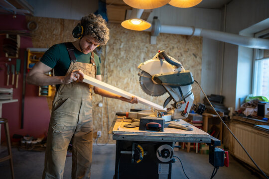 Serious concentrated middle aged carpenter man measures and inspects wooden beam before cutting with mitre saw in workshop. Craftsmanship accuracy for woodworking project, preparation stage - Powered by Adobe