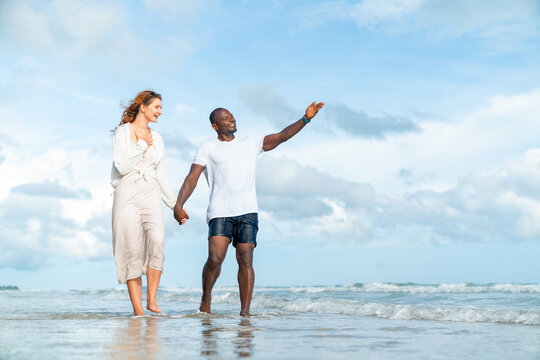 Happy man and woman travel nature the sea on summer beach holiday vacation. Diverse family couple enjoy outdoor active lifestyle walking and relaxing together on tropical island beach at summer sunset