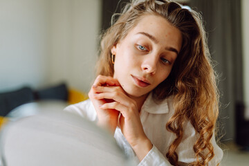 Portrait of a young woman looking into a small mirror and examining skin imperfections in a cozy room. Cute woman touching rashes indoors with hands. Concept of skin care, dermatology.