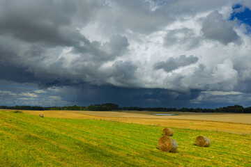A dramatic sky with dark storm clouds looms over golden fields and green meadows dotted with hay bales.