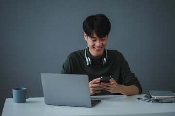 Asian freelancer smiling and using smartphone while working at home with laptop, headphones and coffee mug on white desk