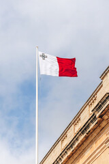 The national flag of Malta with the George Cross flying on a flagpole above a historic building under a cloudy sky.
