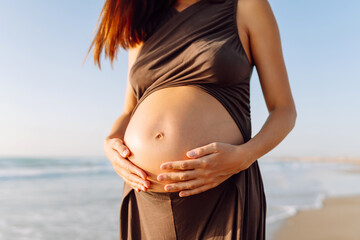 A woman sits on a yoga mat and touches her pregnant belly in the rays of the sunset. Close-up of a young woman practicing yoga and meditation on the seashore. Concept of procreation, health.