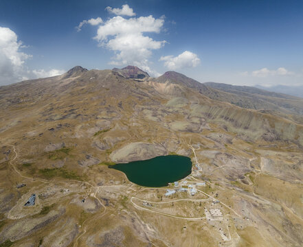 Aerial view of the serene, emerald lake nestled amidst the rugged, sun-drenched slopes of Mount Aragats, under a vast sky dotted with fluffy clouds, Lake Kari, Aragatsotn Province, Armenia.
