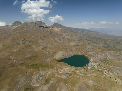 Aerial view of a serene, dark lake mirroring the sky nestled amidst rugged, sun-drenched mountains under a vast, azure sky, Lake Kari, Aragatsotn Province, Armenia.