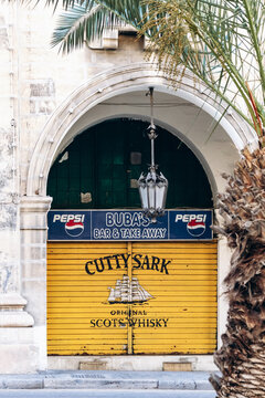 Malta &ndash; April 10, 2025: Closed yellow shutter with &ldquo;Cutty Sark Original Scots Whisky&rdquo; and &ldquo;Buba&rsquo;s Bar and Take Away&rdquo; signage, flanked by archway and palm trees on a quiet street