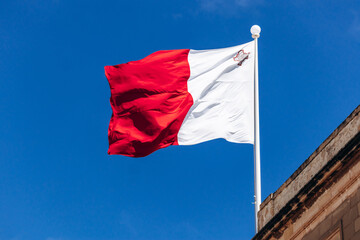 The national flag of Malta, red and white with the George Cross in the upper hoist corner, flies against a clear blue sky above traditional Maltese limestone architecture