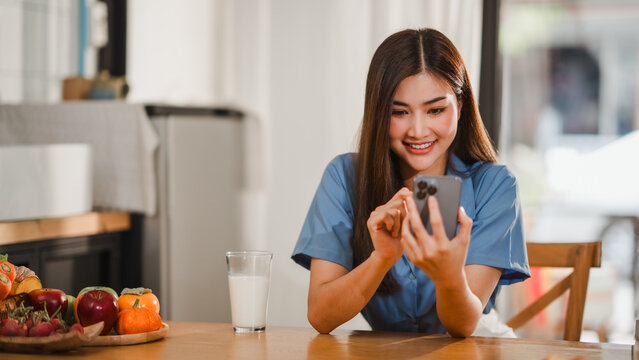 Happy young woman using smartphone while having breakfast in kitchen - Powered by Adobe