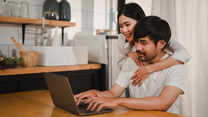 Asian couple using laptop in kitchen, woman hugging man working from home