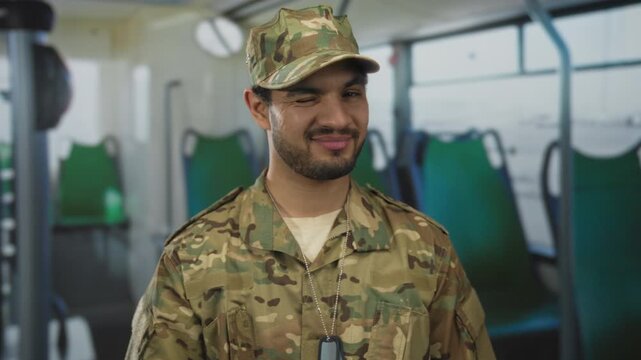 Man in camouflage uniform winks inside bus on street with dogtags around neck by green seats; duty.