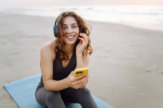 Smiling woman in sportswear with phone and wireless headphones sitting on yoga mat on beach. Young woman enjoying listening to music and seascape after workout. Nature, technology concept.