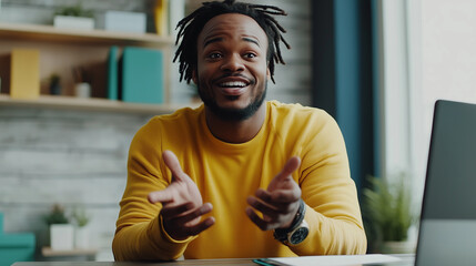 Cheerful young man in yellow sweater is sitting at desk with laptop, gesturing with his hands while engaging in lively conversation in modern home office setting
