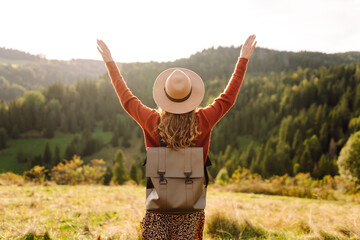 A joyful woman with a backpack and a hat walks through green hills at sunset. A traveler enjoys...