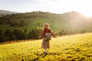 Fototapeta premium A joyful woman with a backpack and a hat walks through green hills at sunset. A traveler enjoys nature in the mountains and feels free. Active lifestyle.