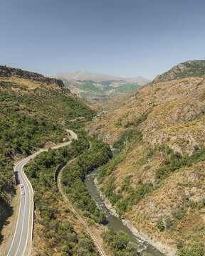 Aerial view of a winding road snaking through a valley alongside a river, framed by rugged, sun-baked hillsides dotted with green vegetation, Lori, Lori Province, Armenia.