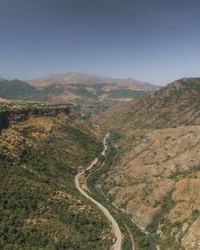 Aerial view of a winding road snaking through the rugged, sun-drenched Debed Canyon, contrasting with the distant peaks under a clear sky, Lori, Lori Province, Armenia.