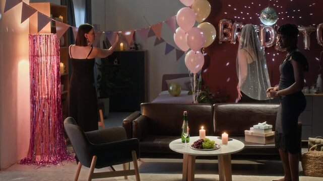 Group of young diverse women in elegant black dresses decorating room and preparing wine glasses, then gathering around table with champagne and candles for festive bachelorette party