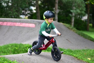 Toddler boy wearing helmet rides red balance bike on asphalt pump track. Active child learning balance and coordination while enjoying outdoor play in park.