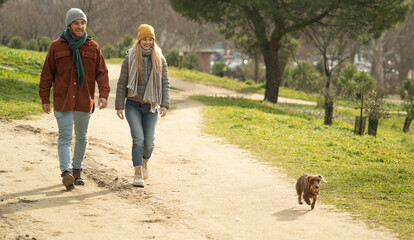 Smiling young couple walking with their dachshund dog in a park, enjoying a sunny winter day