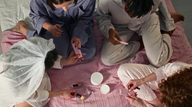 Top view of bride-to-be and girlfriends sharing nail polish, toe separators and facial patches on bed while preparing for skincare routine and pedicure during bachelorette party