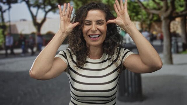 Woman sticks tongue out and raises hands by temples on a sunny city street; playful banter defiance.
