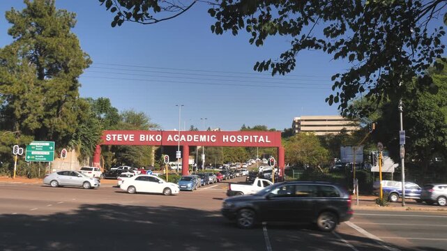 Wide view, many vehicles cross traffic light intersection in slow motion at Steve Biko Academic Hospital main entrance