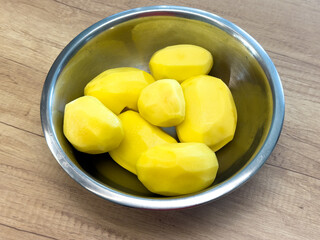 Stainless steel bowl filled with peeled potatoes on wooden table, fresh vegetables prepared for cooking.