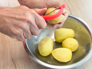 Hands peeling potato with red peeler above metal bowl with peeled potatoes on wooden table, kitchen preparation.