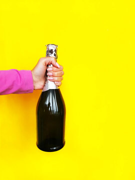 Hand In Pink Clothes Holding a Closed Bottle of Champagne on Yellow Monochrome Background