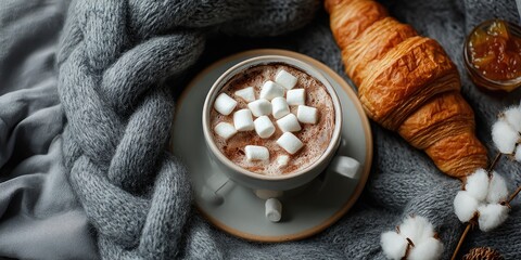 Cozy hot chocolate with a croissant, topped with marshmallows, on a gray knitted blanket