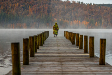 A boy on a Windermere Jetty, wearing a Camo changing robe, an Autumn scene with mist rising from the lake. © Ian