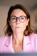 Fototapeta premium A focused woman in a pink blazer, wearing glasses, stares intently at the camera, conveying confidence and professionalism in a soft-lit environment.