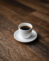 A simple white coffee cup filled with black coffee sits on a white saucer against a rich wooden surface.