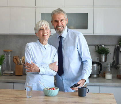 Portrait of a mid aged mature woman and man  gettyng ready for work using a smartphone and eating breakfast oatmeal in a bowl standing in kitchen home. Overworked, busy, stress, urgency, multitasking,