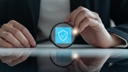 Businessman holding a magnifying glass with a digital shield and a checkmark icon, cybersecurity,...