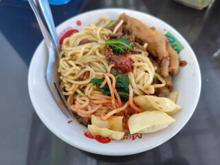 A complete bowl of chicken noodles with claws, dumplings, chicken pieces and red chili sauce on top, traditional Indonesian food.