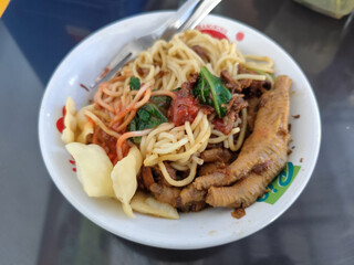 A complete bowl of chicken noodles with claws, dumplings, chicken pieces and red chili sauce on top, traditional Indonesian food.
