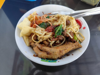 A complete bowl of chicken noodles with claws, dumplings, chicken pieces and red chili sauce on top, traditional Indonesian food.