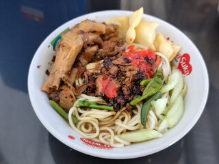 A complete bowl of chicken noodles with claws, dumplings, chicken pieces and red chili sauce on top, traditional Indonesian food.