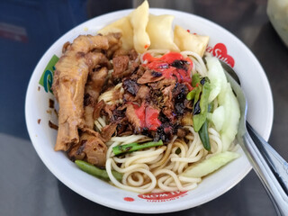 A complete bowl of chicken noodles with claws, dumplings, chicken pieces and red chili sauce on top, traditional Indonesian food.