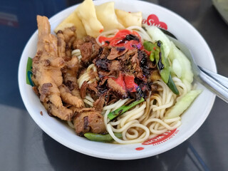 A complete bowl of chicken noodles with claws, dumplings, chicken pieces and red chili sauce on top, traditional Indonesian food.