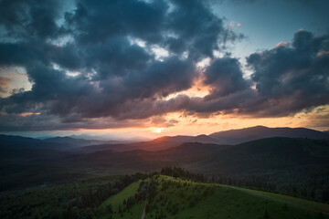 Aerial view breathtaking sunset over forest. Sun's rays break through dramatic clouds, casting golden glow across rolling hills and valleys. Distant mountains creating serene and picturesque scene.