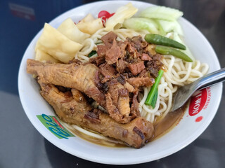 A complete bowl of chicken noodles with claws, dumplings, chicken pieces and red chili sauce on top, traditional Indonesian food.