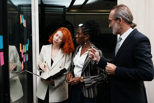group of employees and an elderly male executive and two multinational female team members stand together, brainstorming and debating a strategic plan using stickers on a glass wall in conference room