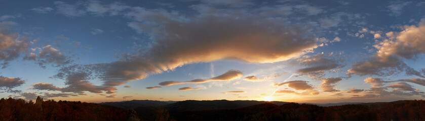 Obraz premium Stunning sunset fills sky with rich hues of blue and orange. Dramatic clouds with hints of pink and gold spread across sky. Silhouettes of hills in foreground add depth to breathtaking evening scene.