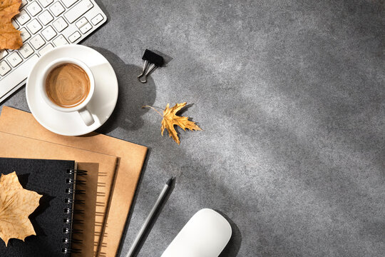 An autumn desktop with a keyboard, coffee cup, notebooks, mouse, pencil, binder clip and scattered fall leaves on a gray desk surface.