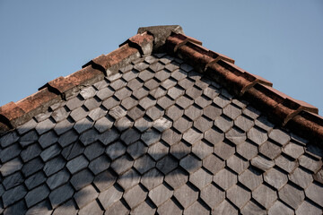 A traditional roof peak with gray hexagonal wooden shingles and weathered clay ridge tiles against a clear blue sky. An architectural detail background.