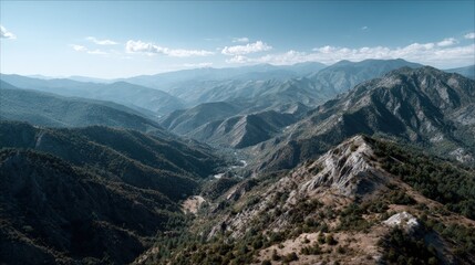 Naklejka premium A sweeping vista showcases a mountain range, with deep valleys and lush greenery, under a clear blue sky dotted with fluffy clouds.