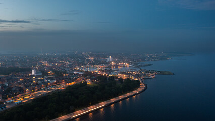 Aerial night view of Thisted, Denmark. Town lights, marina, Limfjord coastline, and surrounding neighborhoods create a tranquil Nordic cityscape under a twilight sky